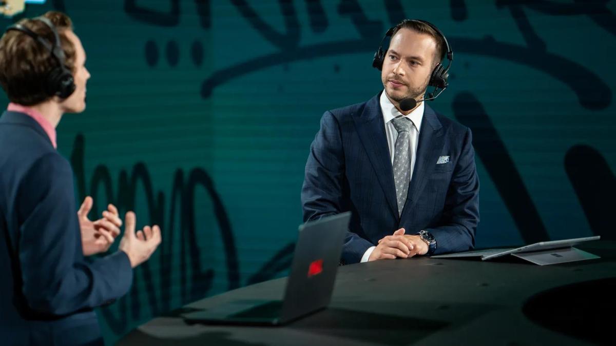 Counter-Strike personality Maniac watches another CS2 talent speak to him with a neutral look on his face while leaning on an ESL stage desk.