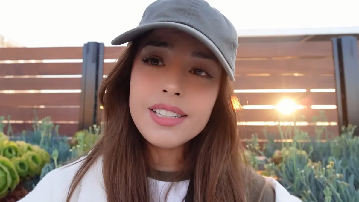 Valkyrae wearing a hat and sitting in front of a camera on a balcony.